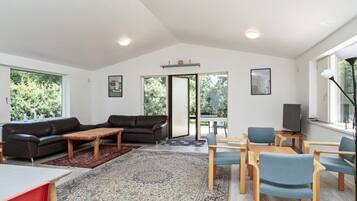A modern living room with a sofa, coffee table, and chairs, a TV, and a large window with a view of trees.