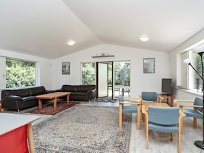 A modern living room with a sofa, coffee table, and chairs, a TV, and a large window with a view of trees.