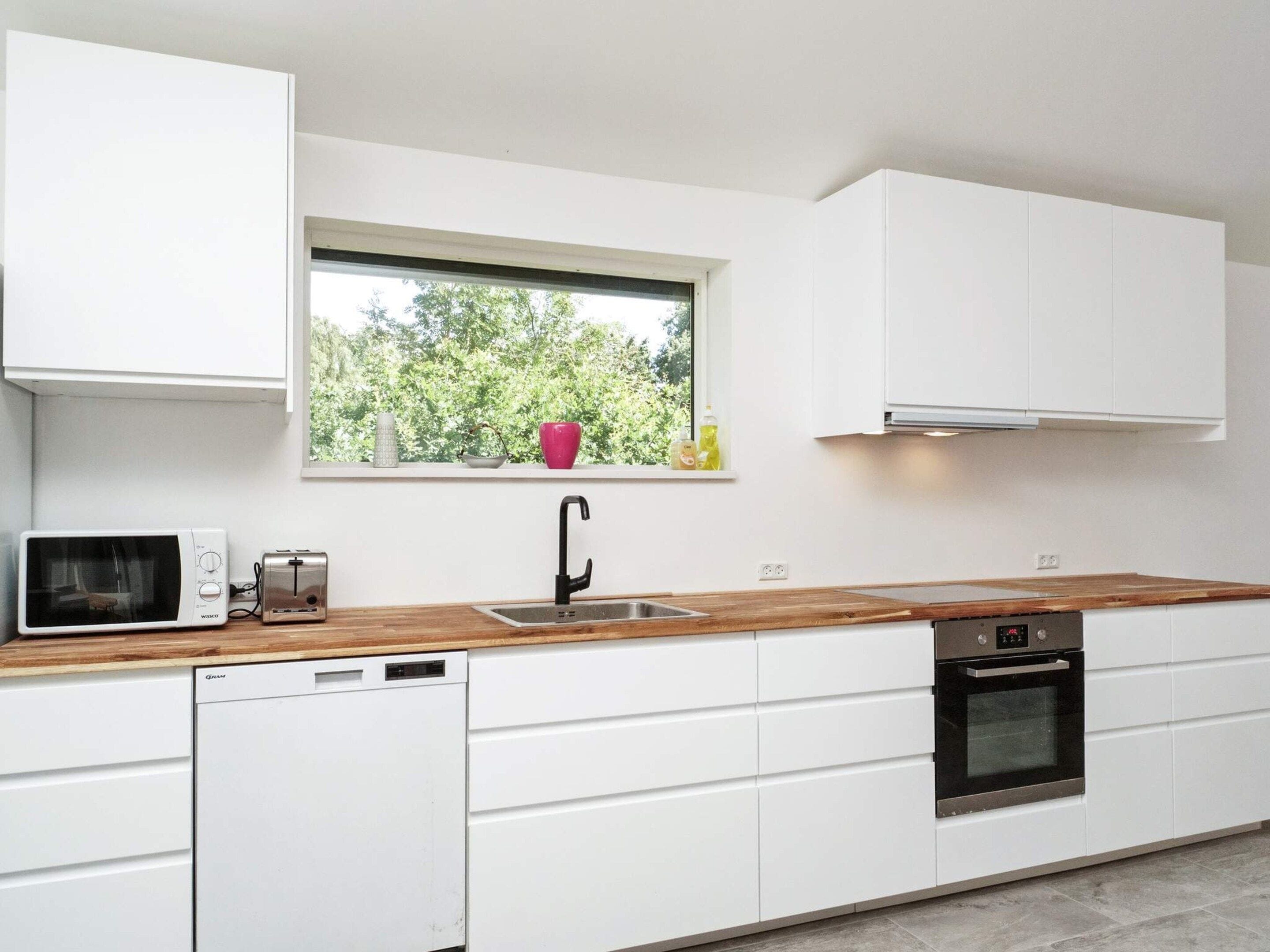 A modern kitchen with white cabinets, a wooden countertop, a built-in dishwasher, a stainless steel oven, and a window with a view of trees.