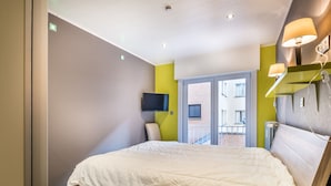 A modern hotel room with a large bed, a flat-screen TV mounted on the wall, and a balcony with a view of the city. - Apartment Near Dunes With Sea View (Koksijde)