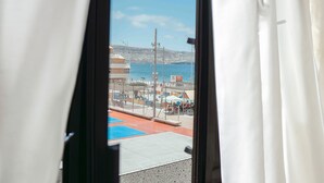 A view through a window with white curtains, revealing a beachside area with a blue and red floor, a fence, and a clear sky.