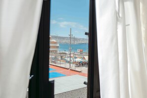 Blick durch ein Fenster mit weißen Vorhängen auf eine Strandlage mit blauem und rotem Boden, einem Zaun und einem wolkenlosen Himmel.