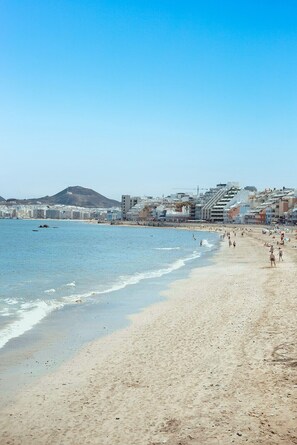 Ein Strand mit einer Stadtlandschaft im Hintergrund, darunter Gebäude und ein Hügel.