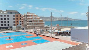 A sports court with people playing, a beach with people and umbrellas, and buildings in the background.