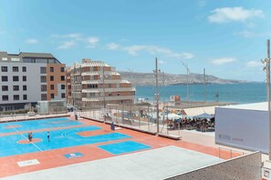 Ein Sportplatz mit spielenden Personen, ein Strand mit Menschen und Sonnenschirmen sowie Gebäude im Hintergrund.