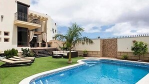 A swimming pool with a tiled edge, surrounded by a grassy area, a stone wall, and a building with a balcony. - Belvilla by OYO Villa in Adeje (Adeje)