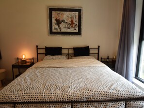 A neatly made double bed with a patterned bedspread, two black pillows, a framed artwork above the bed, a bedside table with a lamp, and a window with curtains. - Modern Holiday Home in Maasmechelen With Sauna (Maasmechelen)