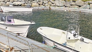 Un puerto deportivo con varias embarcaciones amarradas, una furgoneta blanca estacionada cerca y una costa rocosa al fondo.