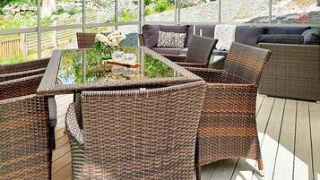 An outdoor patio area with wicker furniture, a glass-top table, and a view of a garden and stone wall.