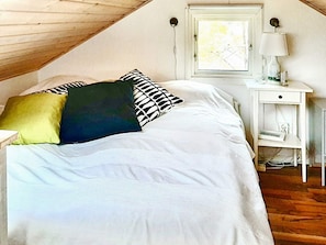 Een slaapkamer met een schuine kap, een bed met witte beddengoed, een nachtkastje met een lamp en een raam met uitzicht.