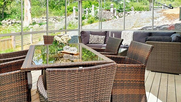 Un espace extérieur aménagé avec des meubles en osier, comprenant une table à plateau de verre et des chaises rembourrées, offrant une vue sur un jardin et un sentier pavé de pierres.