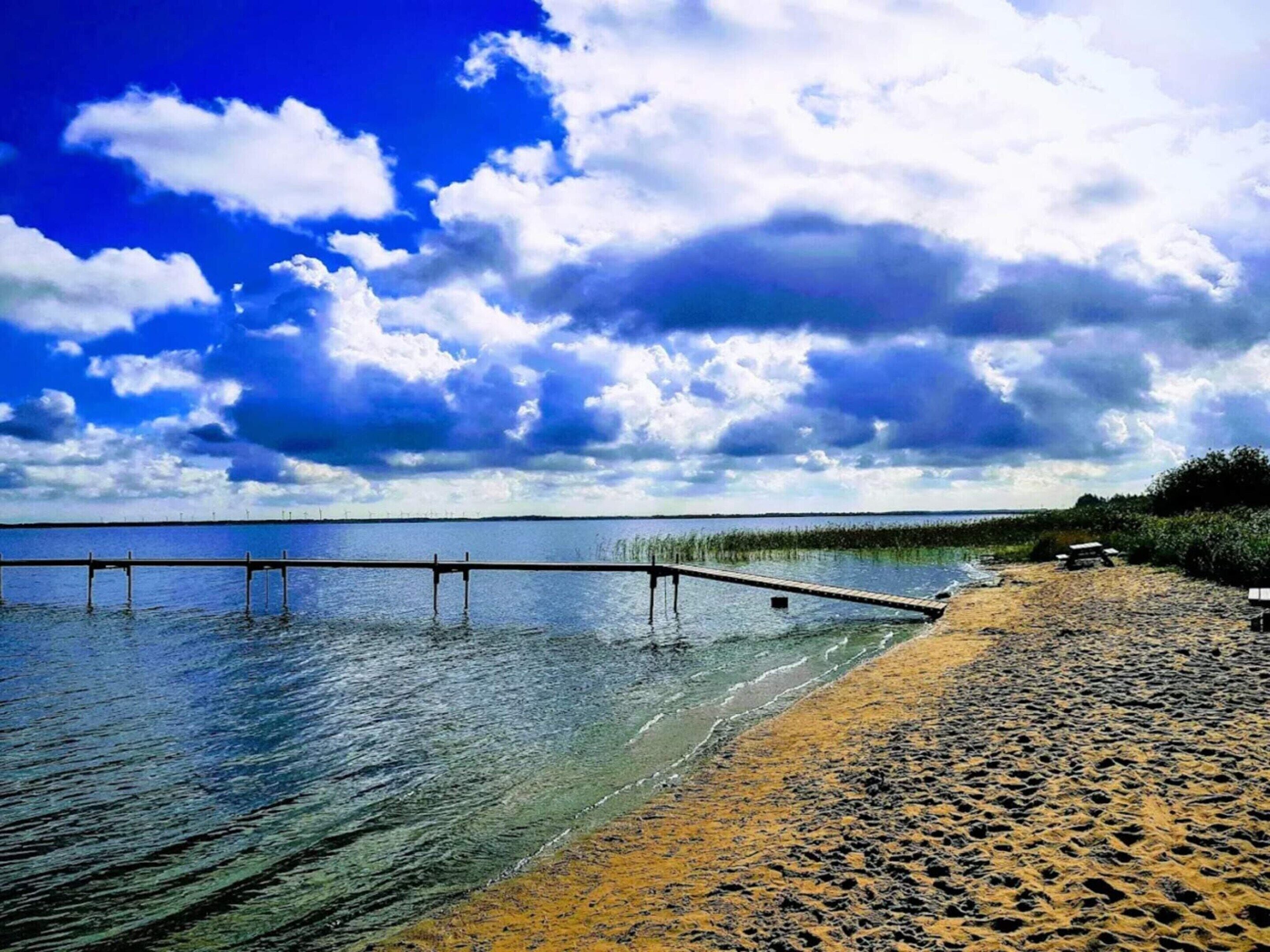 Une plage avec une jetée en bois qui s’avance dans des eaux calmes, le tout baigné d’un ciel dégagé.