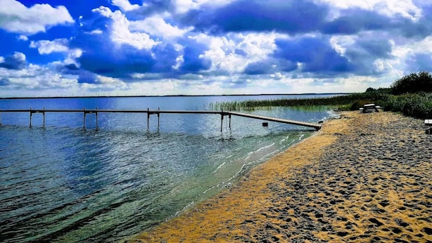 Una playa con un muelle de madera que se adentra en aguas tranquilas, rodeada por un cielo despejado.