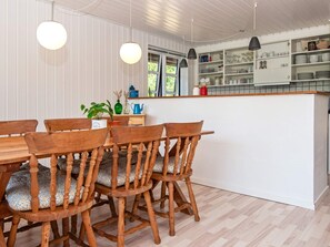 Un comedor con sillas de madera y una mesa, una cocina con armarios y estantes blancos, e iluminación colgante.