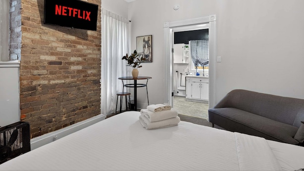 Ein Hotelzimmer mit Backsteinwand, einem Bett mit weißen Leinentextilien, einem grauen Sofa, einem kleinen Tisch mit einer Vase und einem Fernseher, der 'NETFLIX' anzeigt.