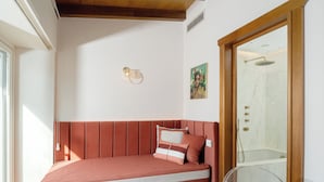 Une pièce avec un canapé-lit rouge, une petite table sur laquelle se trouve un livre, un luminaire fixé au mur et un espace douche visible à travers une porte en verre.
