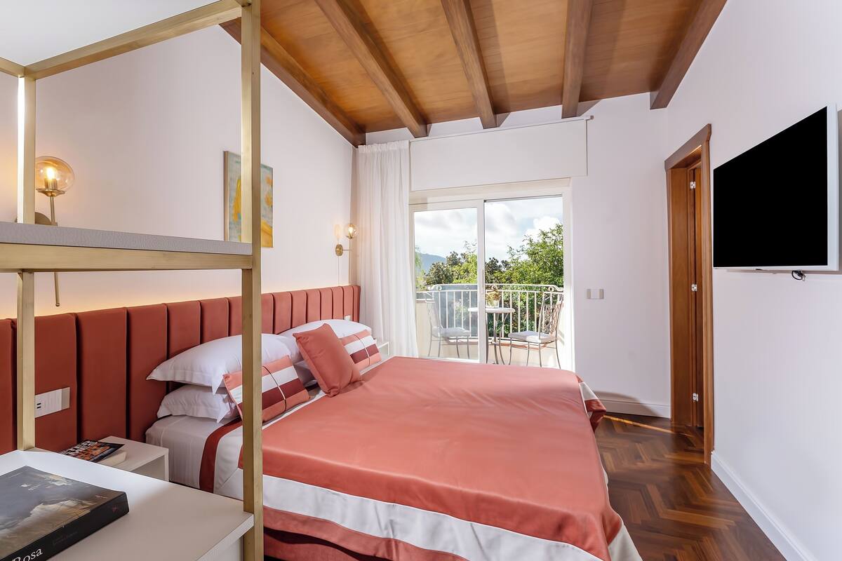 Habitación de hotel con techo de madera, una cama con colcha roja y blanca, una mesita de noche con un libro y un televisor montado en la pared.