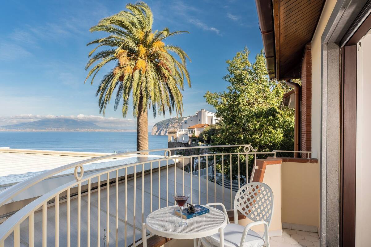 Un balcón con una mesa y sillas blancas, una palmera y vista a la playa.