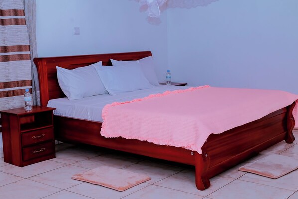 Una cama bien hecha con sábanas blancas y una manta rosa, una mesita de noche de madera con una botella de agua, y un suelo de baldosas.