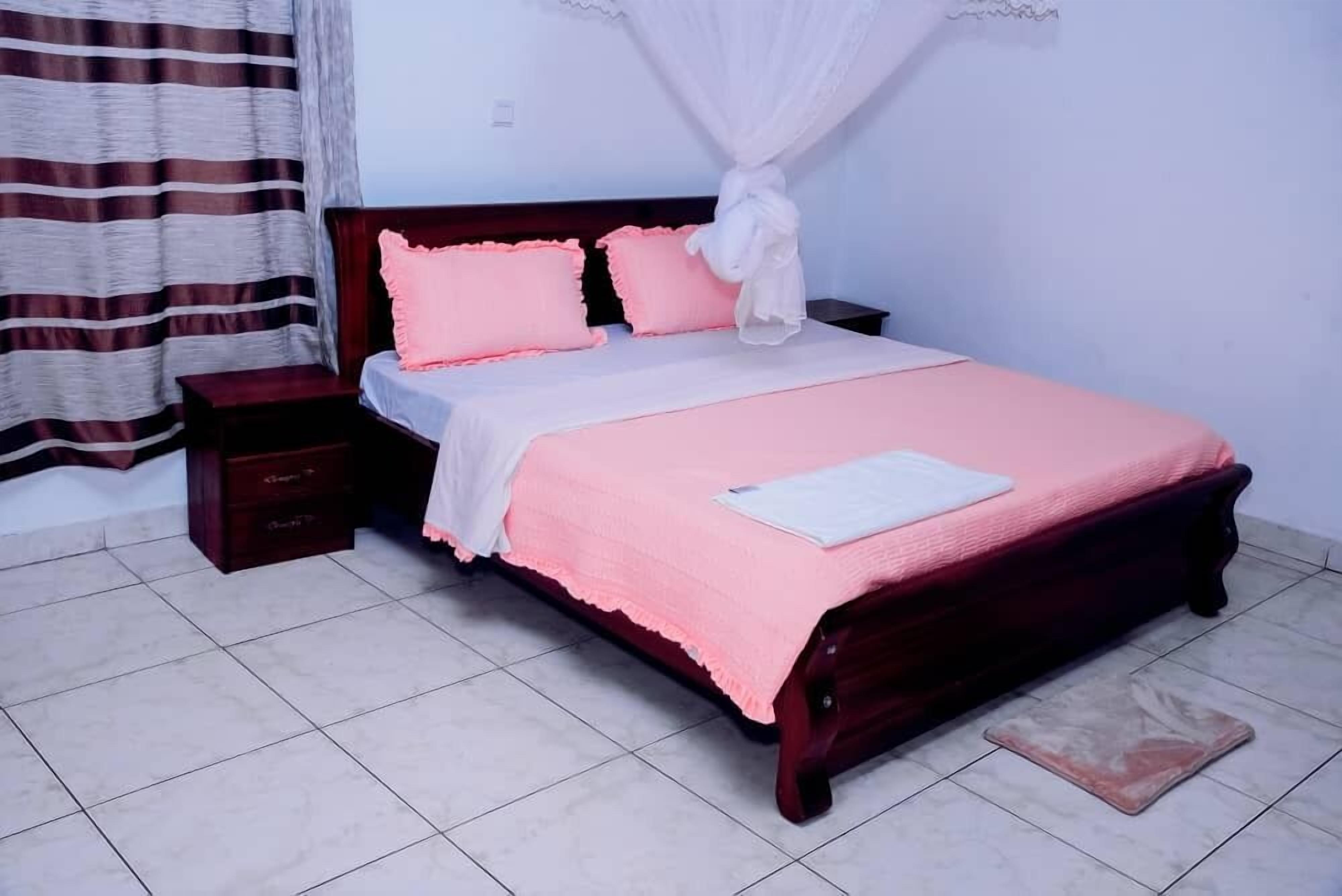 A neatly made bed with pink bedding and a white pillow, a wooden nightstand with drawers, and a curtain with a striped pattern.