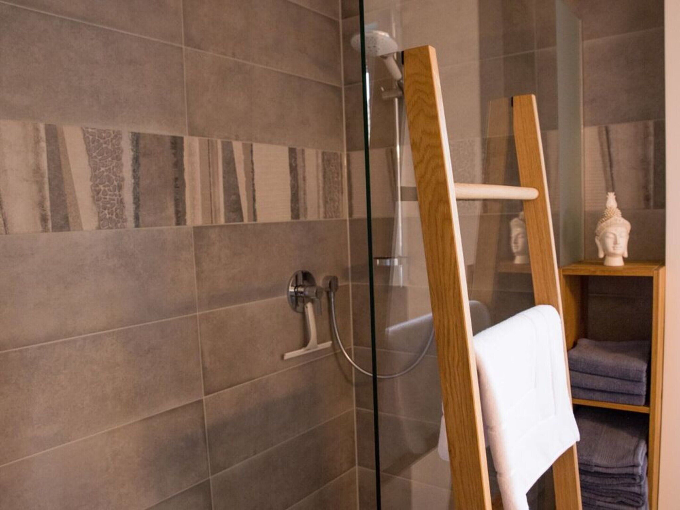A modern bathroom with a glass shower enclosure, a wooden towel rack, and a shelf with folded towels.