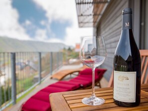 A bottle of Muller Dornfelder wine and an empty glass on a wooden table on an outdoor balcony with red cushions. - Emerald - Holiday Home for (Burgen)