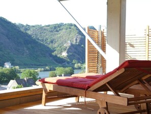 A wooden deck with a red lounge chair overlooking a river and hills. - Emerald - Holiday Home for (Burgen)