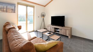 En moderne stue med en brun lædersofa, et fladskærms TV på et træstel, et lille bord og en gulvlampe.