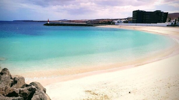 A coastal beach with clear turquoise waters, a sandy shore, and a lighthouse in the distance. - Apartment in Puerto del Rosario Near Playa Chica (Puerto del Rosario)