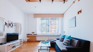 A modern living room with a large flat-screen TV, a dark-colored sofa with blue cushions, a glass coffee table, and a wooden ceiling structure.