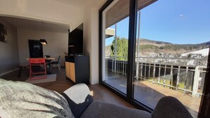 A modern living room with a large window, a sofa, a TV, and a small dining area. - Villa in Winterberg Near the ski Area (Winterberg)
