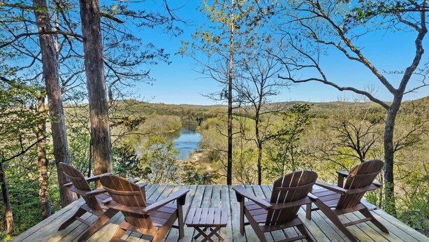 A wooden deck with four Adirondack chairs and a small table, overlooking a river and forested landscape. - Sycamore Ranch - Private MTB Trails - Hot tub! (Eureka Springs)