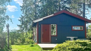 Pieni, yhden kerroksen mökki, jossa on punainen ovi ja ikkuna. Sen ympärillä on vehreää kasvillisuutta ja korkeita puita.