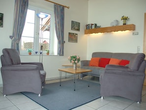 Ein Wohnzimmer mit zwei grauen Sesseln, einem grauen Sofa mit roten Kissen, einem Holztisch und einem blauen Teppich. Zwei Fenster mit Vorhängen, eine Stehlampe und ein Regal mit Dekorationen sind ebenfalls vorhanden.