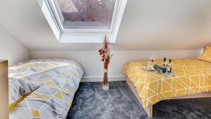 A small, modern bedroom with a skylight, two beds with geometric-patterned bedding, a vase with dried flowers, and a tray with bottles and snacks on one bed. - Bridge House in Skinningrove (Saltburn-by-the-Sea)