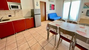 A compact kitchen with red cabinets, a microwave, and a refrigerator. There is a dining table with chairs and a blue sofa in the living area. - Relaxing Apartment in Caorle for 7 (Caorle)