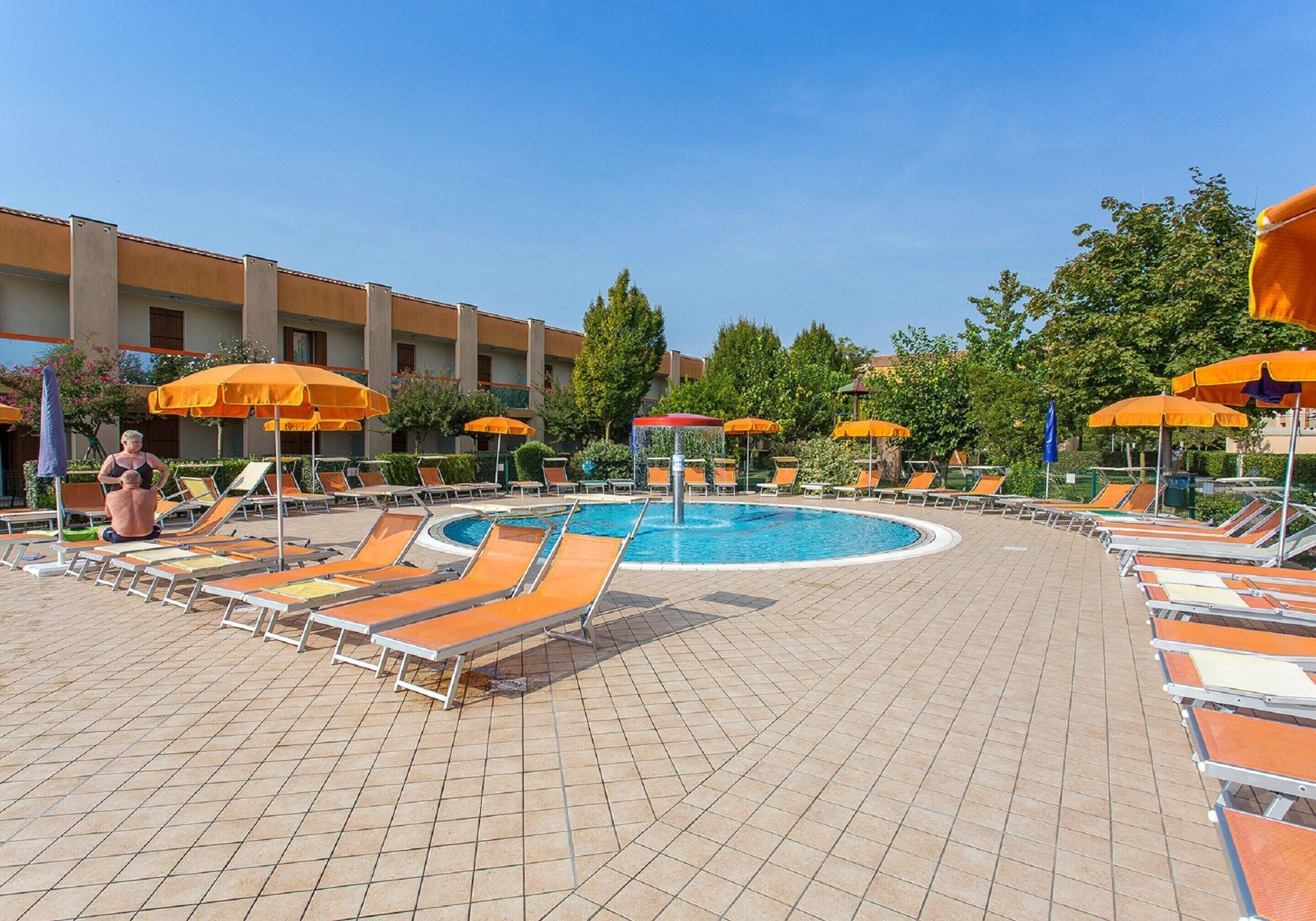 A pool area with sun loungers and umbrellas, a central water feature, and a building with multiple windows in the background.