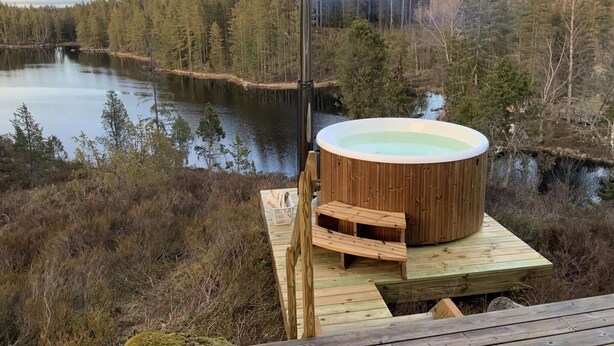 A hot tub on a wooden deck overlooking a lake surrounded by trees. - Holiday Home by the Lake (Tived)