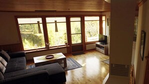 A living room with a sofa, a coffee table, and a television. There are large windows providing natural light and a view of greenery outside. - Holiday Home by the Lake (Tived)