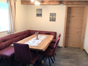 Un coin repas avec une table en bois, des chaises à revêtement rouge et un chemin de table à motifs floraux. Deux tableaux encadrés ornent le mur, et une porte en bois est visible au fond.