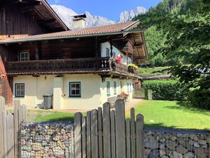 Un chalet traditionnel en bois, doté d’un balcon, avec un mur de pierre au premier plan et une chaîne de montagnes en arrière-plan.