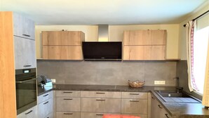 Une cuisine compacte avec des meubles en bois, un four encastré, un micro-ondes, un téléviseur placé au-dessus des meubles et une petite table avec une chaise.