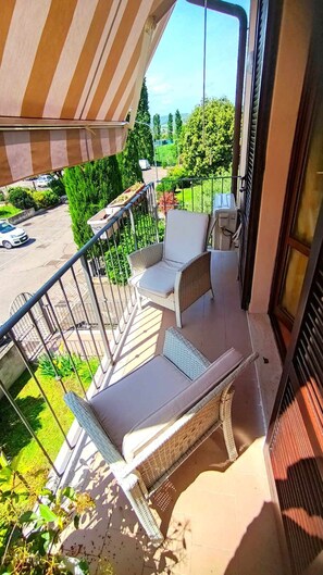 A balcony with two wicker chairs and a small table, overlooking a green landscape and a parking area.
