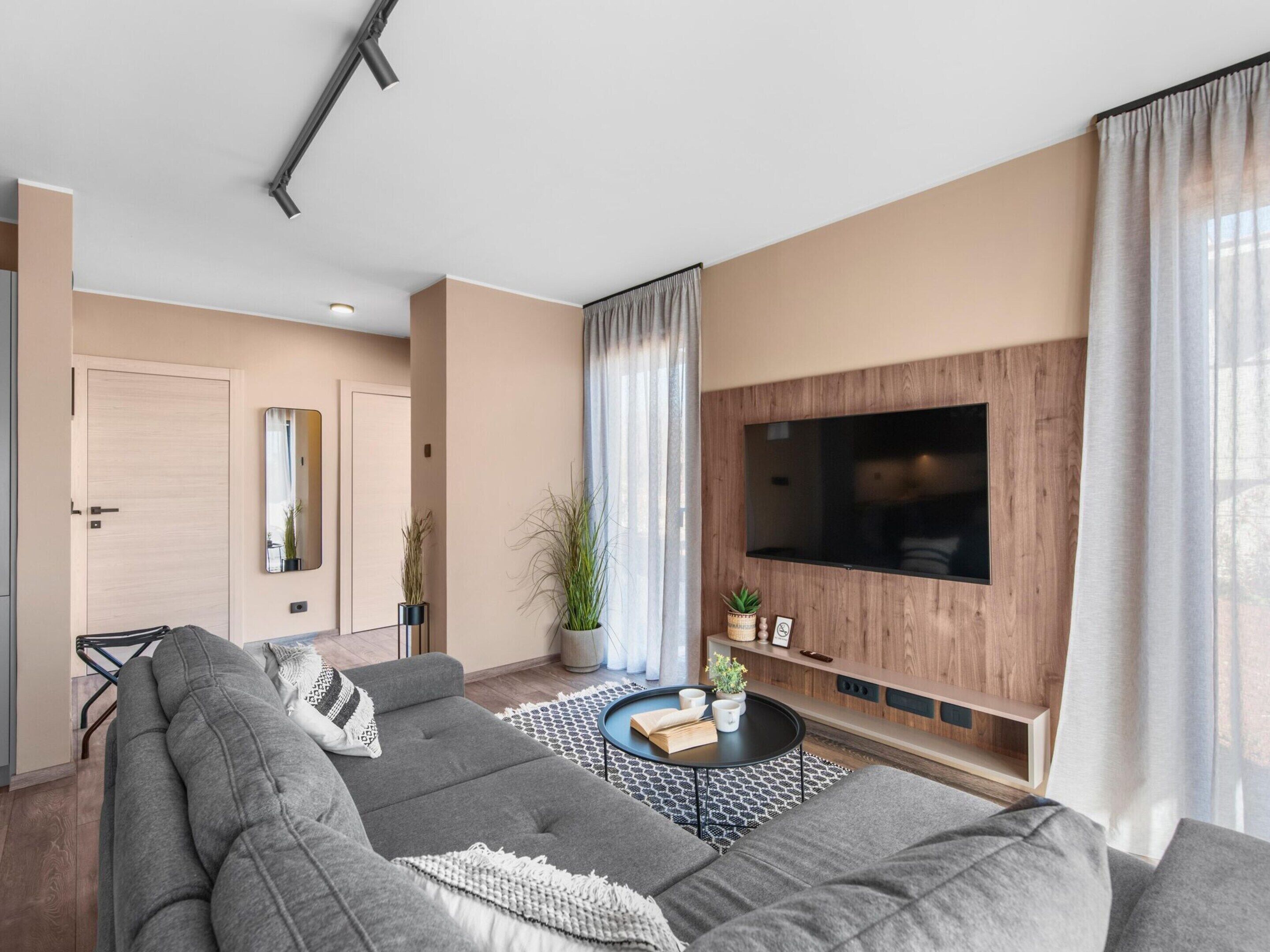 A modern living room with a large sectional sofa, a flat-screen TV mounted on a wooden wall, and a small coffee table with a book and a plant.