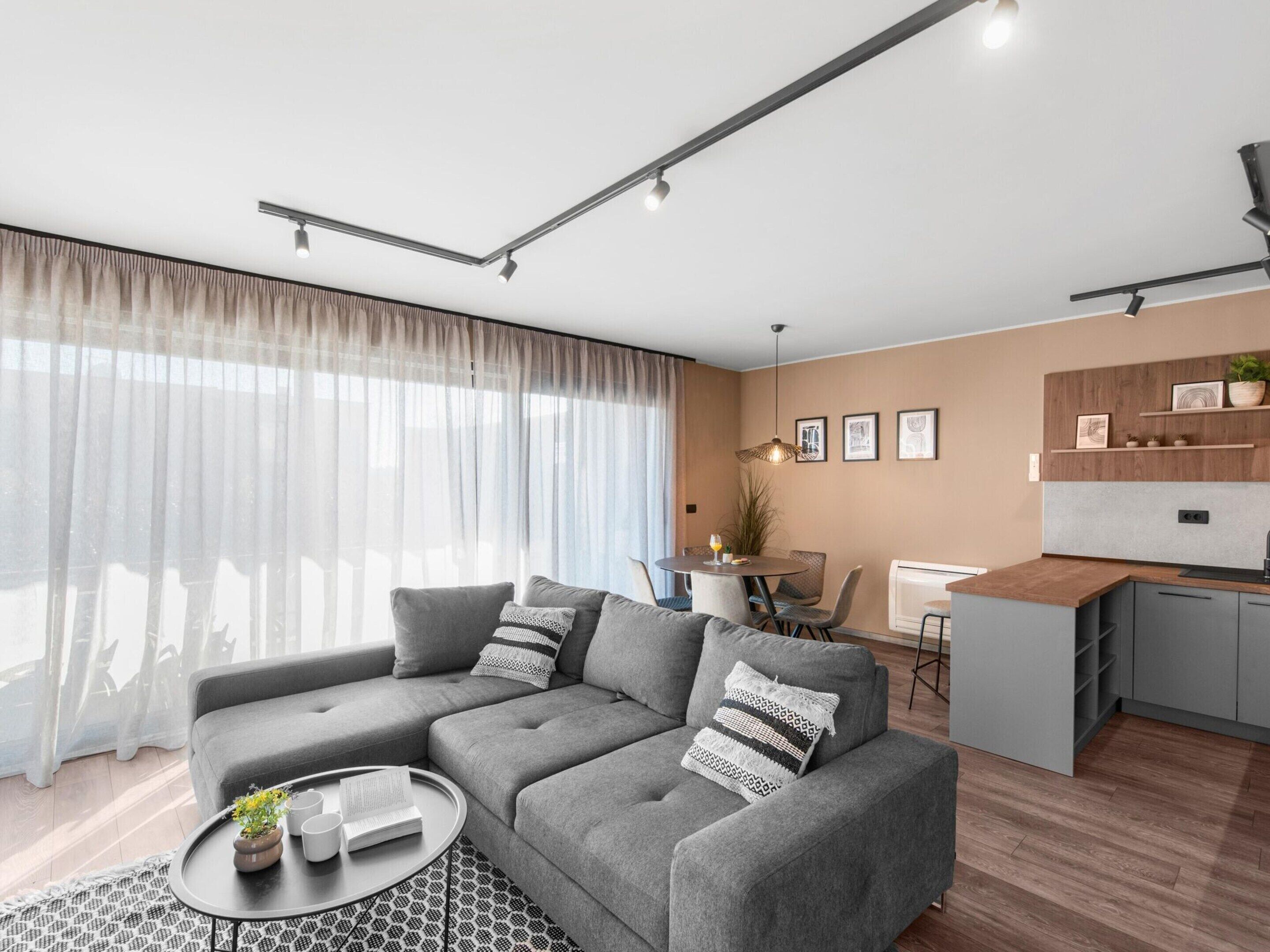 Ein modernes Wohnzimmer mit einem grauen Ecksofa, einem runden Couchtisch und einem Essbereich mit kleiner Küche.