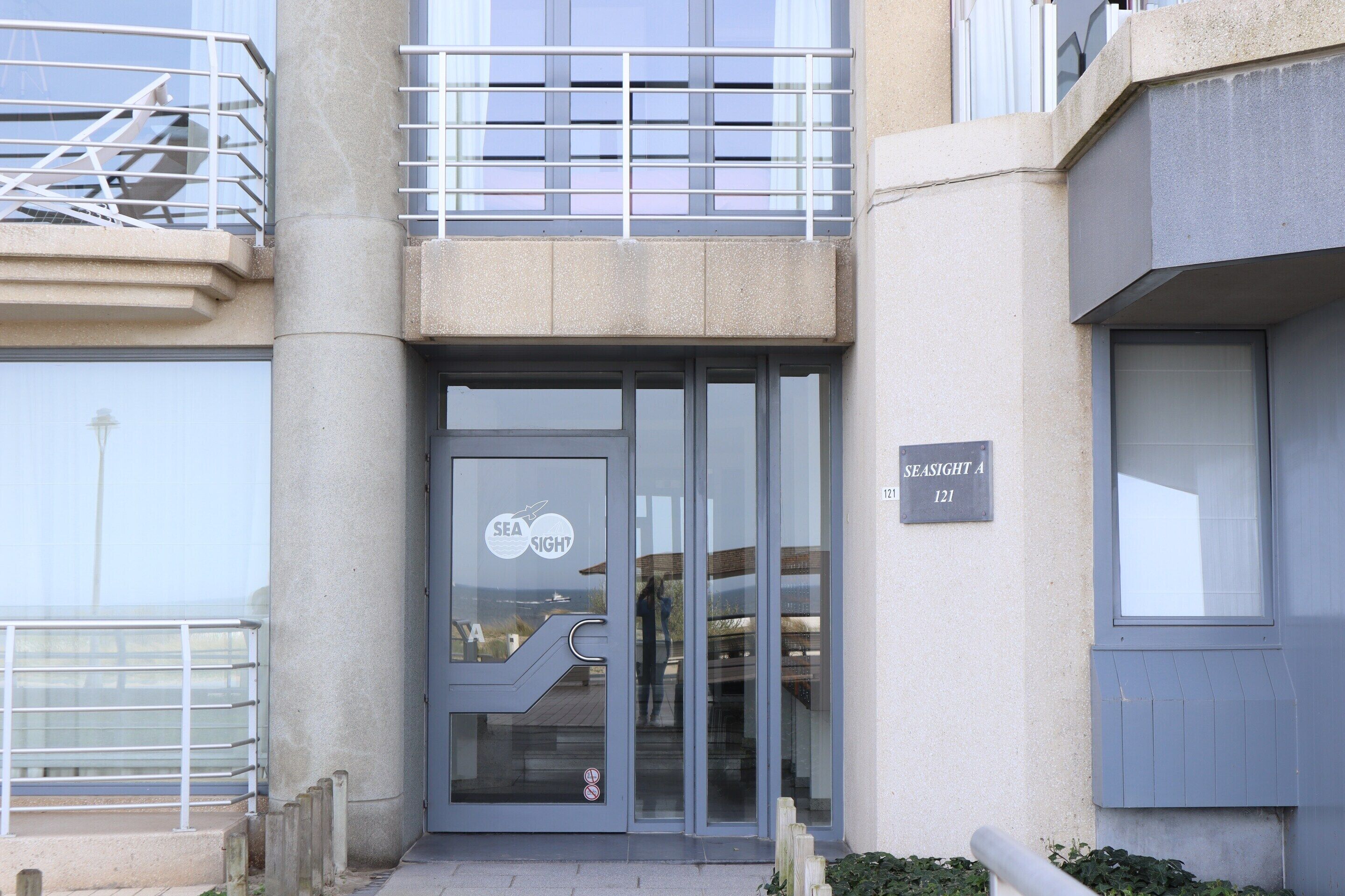 A building entrance with a glass door, a metal railing, and a sign indicating the name and address.