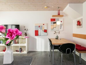 Ett modernt matområde med ett träbord, svarta stolar och en röd kudde. På väggen hänger abstrakta målningar och på en hylla står en vit vas med blommor.