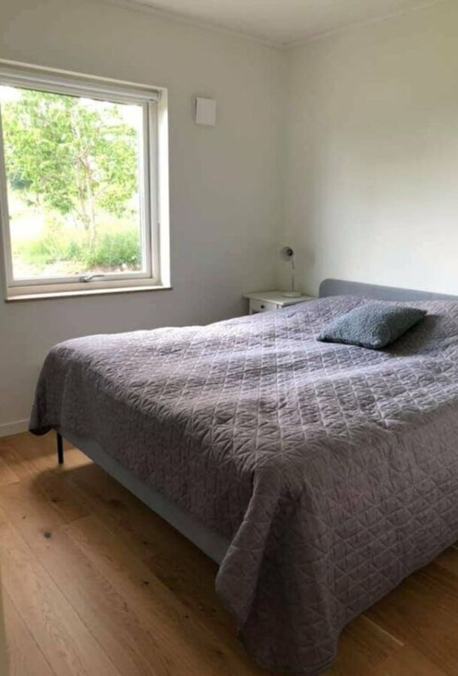 A bedroom with a bed, a nightstand, a window, and a view of greenery outside.