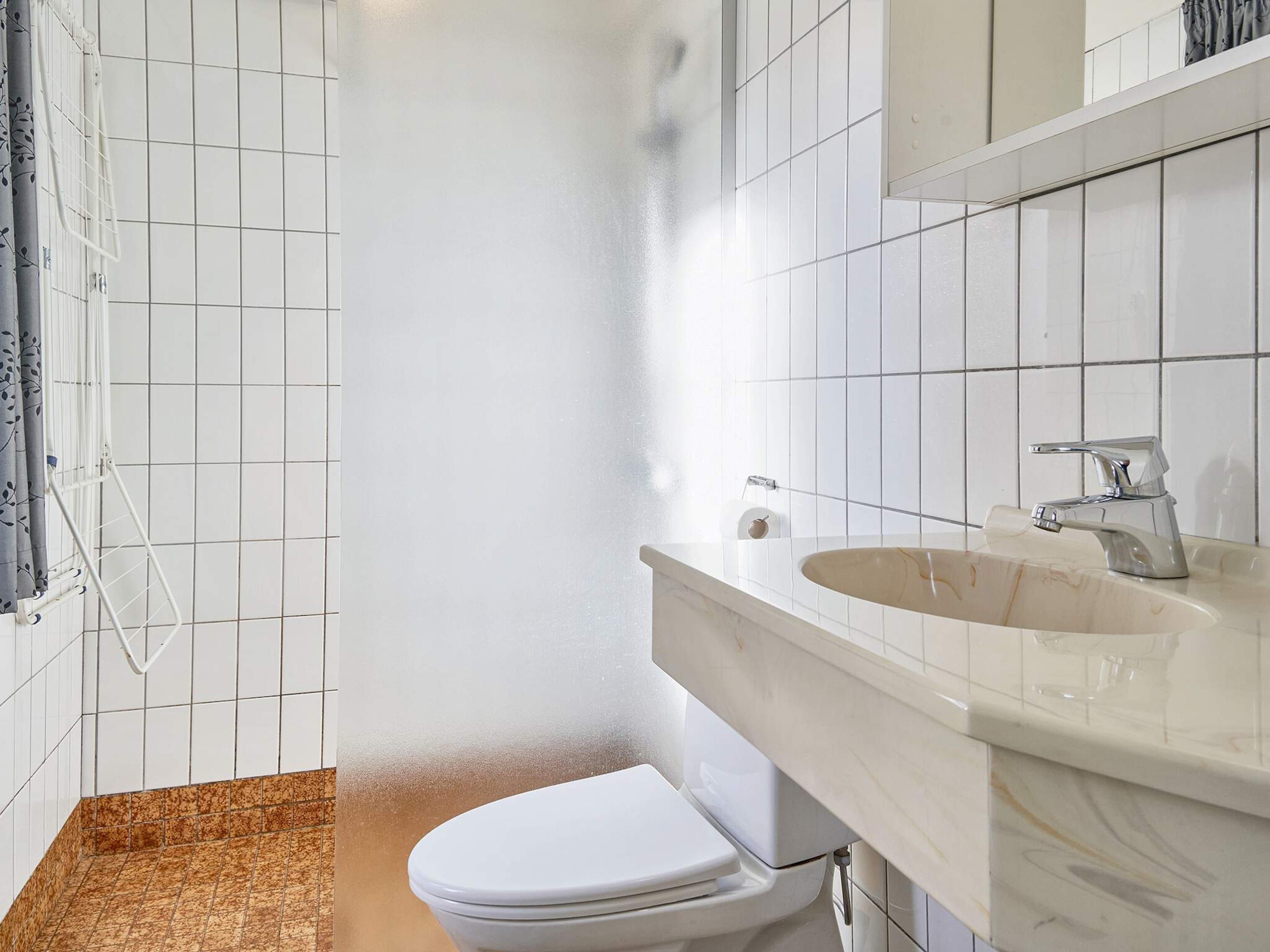 A bathroom with white tiled walls, a white toilet, a beige sink with a modern faucet, and a shower area with a glass door.