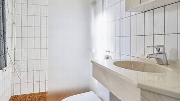 A bathroom with white tiled walls, a white toilet, a beige sink with a modern faucet, and a shower area with a glass door.